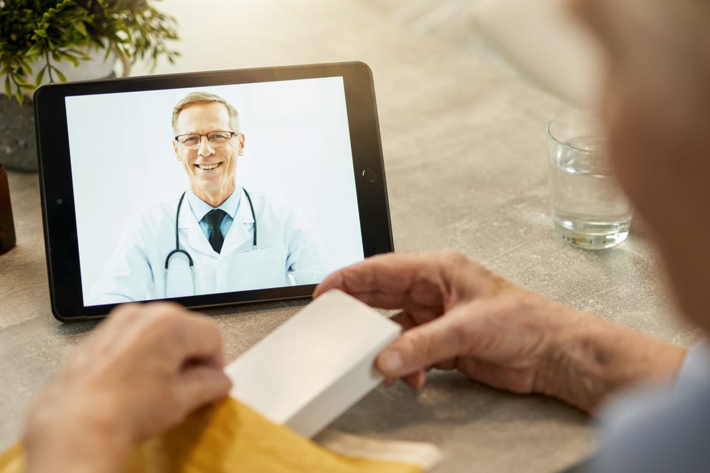 Remote online consultation from a friendly doctor Outgoing medical worker smiling from tablet screen on a video call while his patient holding a pack of pills