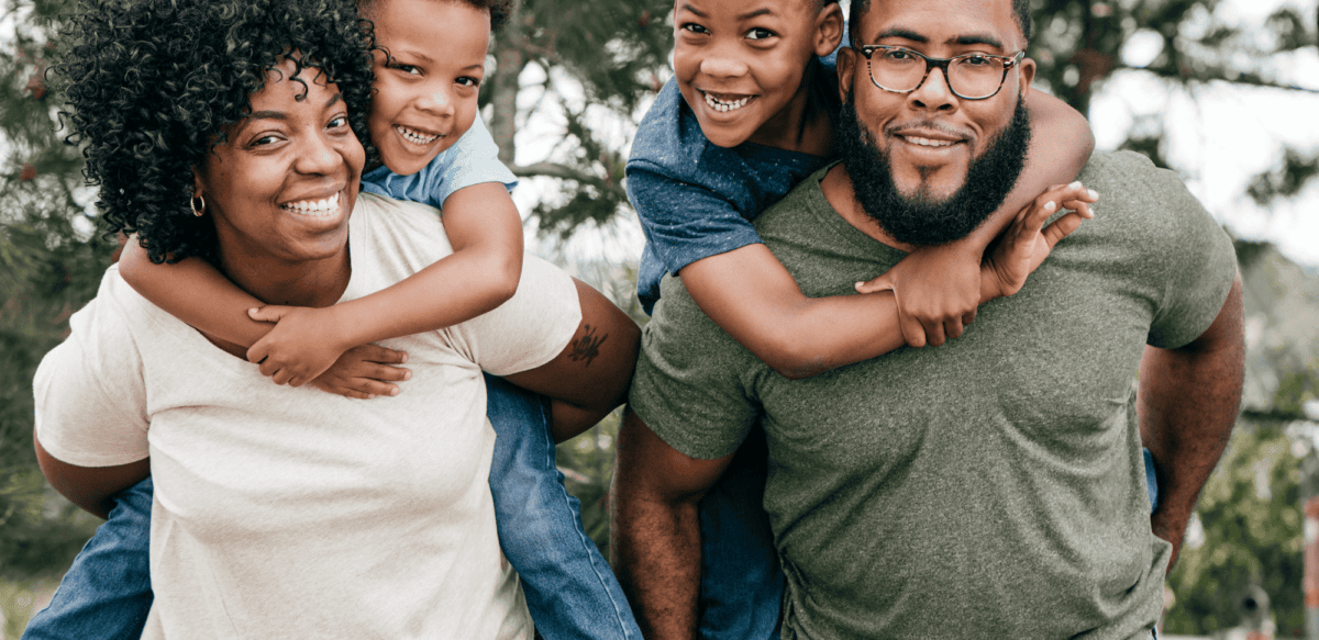 A black mother and father holding two sons on their backs, with trees in the background